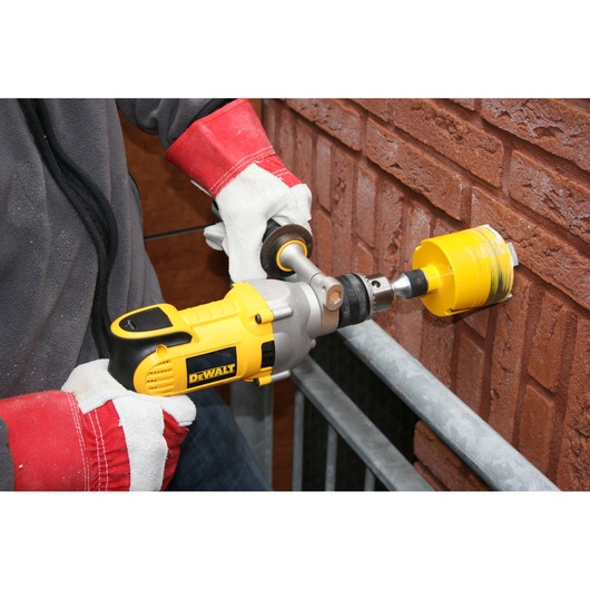 A person wearing protective gloves using a DEWALT D21570K drill with a hole saw attachment to drill into a brick wall.
