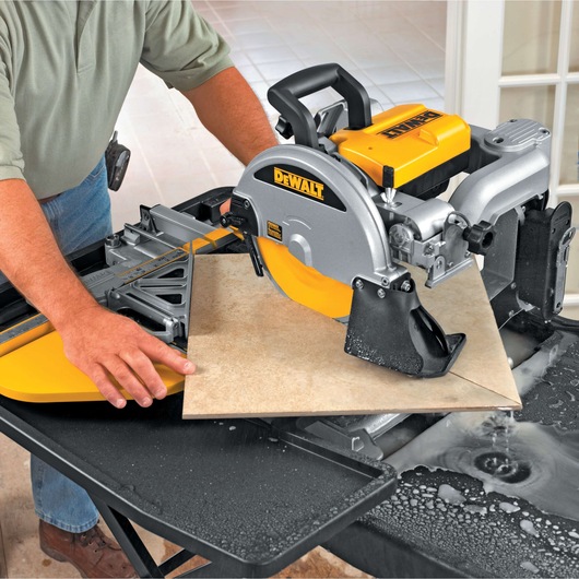 A person using a DEWALT D24000 tile saw to cut a ceramic tile on a worktable, with water helping to cool the blade and reduce dust.
