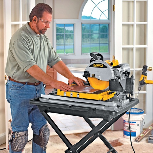 A person wearing knee pads is using a DEWALT tile saw (model D24000) on a stand in a well-lit room with large windows. Construction materials and tools are visible in the background.