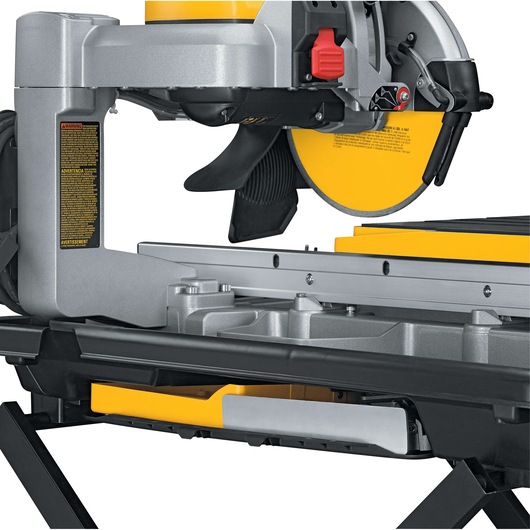 Close-up view of a DEWALT D24000 wet tile saw with a yellow circular blade, mounted on a sturdy metal frame. The saw features adjustment knobs, warning labels, and a built-in water tray for cooling during cutting operations.