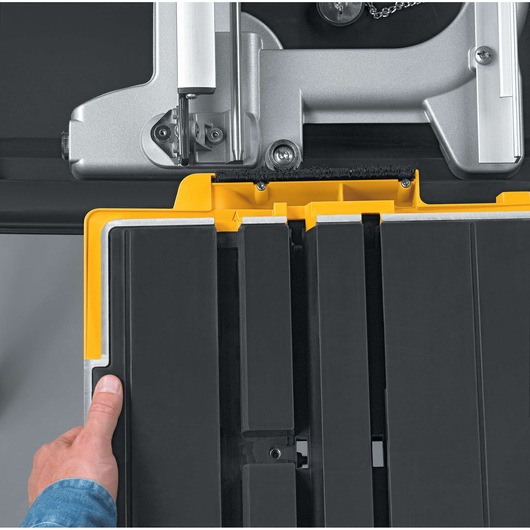 Close-up of a DEWALT D24000 tile saw with a person's hand adjusting the black and yellow sliding tray beneath the cutting blade.