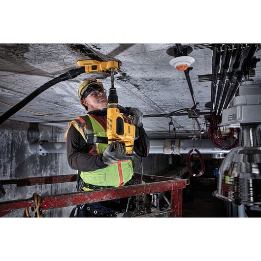 A construction worker in safety gear is using a yellow DEWALT rotary hammer drill to drill into a concrete ceiling. The