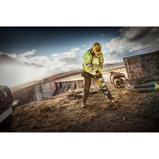 A construction worker wearing a yellow hard hat and high-visibility jacket uses a DEWALT jackhammer on rocky ground near an industrial structure outdoors, with equipment and vehicles visible around.