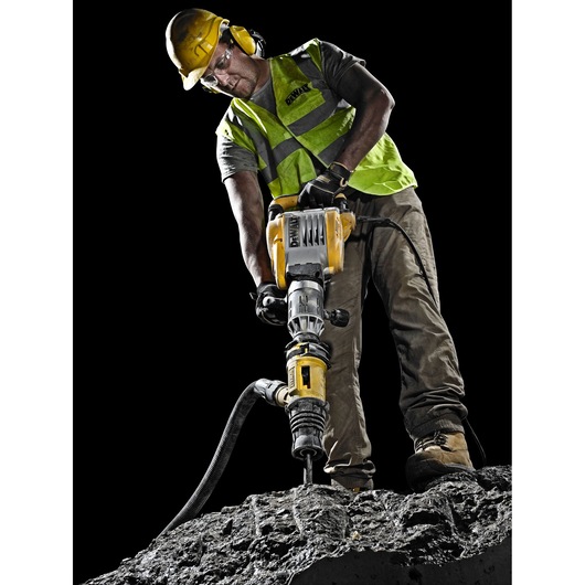 A construction worker wearing a yellow helmet and a high-visibility vest operates a DEWALT demolition hammer on a rough concrete surface. The worker's face is blurred for privacy, and the background is black, highlighting the tool and worker.