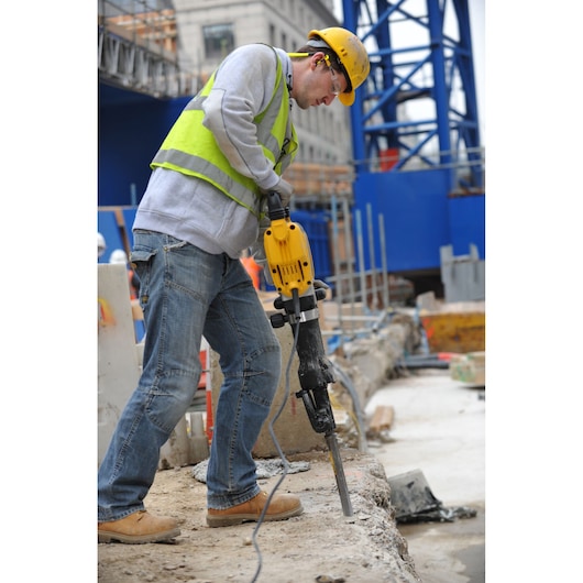 A construction worker using a yellow DEWALT demolition hammer (model D25960K) to break concrete at a building site. The worker is wearing a safety vest, helmet, and boots.
