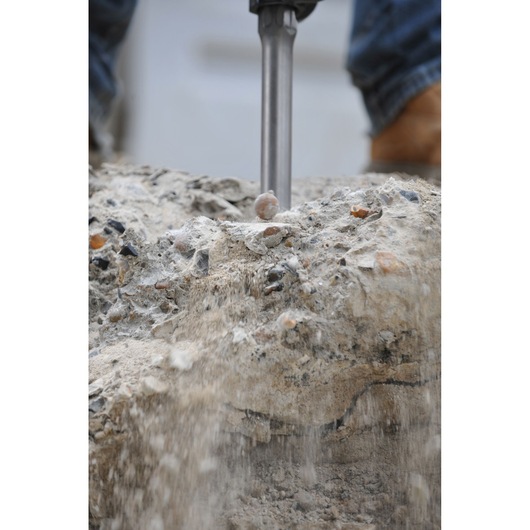 Close-up of a demolition tool breaking concrete, with dust and debris falling, and a person's feet visible in the background.