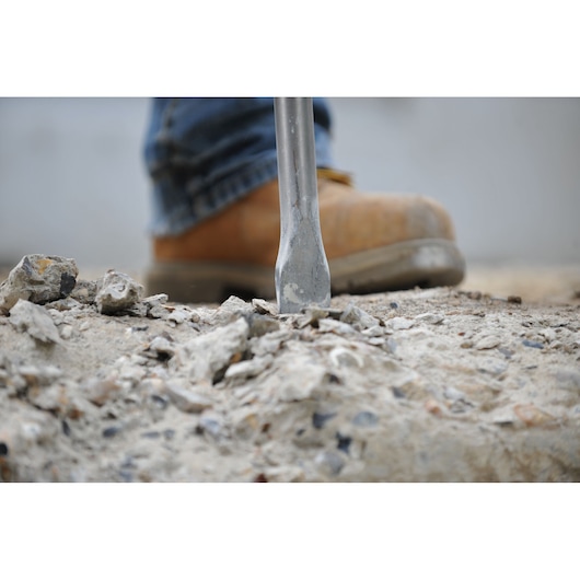 Close-up of a chisel tip breaking concrete, with a worker wearing a brown safety boot in the background.