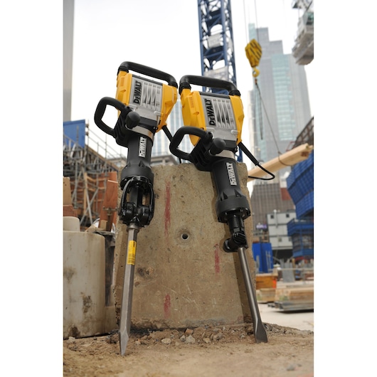Two DEWALT demolition hammers (model D25960K) placed upright against a concrete block at a construction site, with buildings and cranes visible in the background.