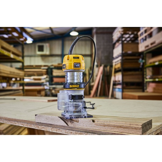 A DEWALT D26200 compact router placed on a wooden board in a workshop, surrounded by shelves and stacks of wood.