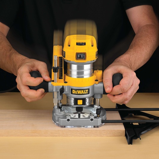 A person using a DEWALT D26203 plunge router to work on a piece of wood. The router is yellow and gray and is held with both hands for precise control.