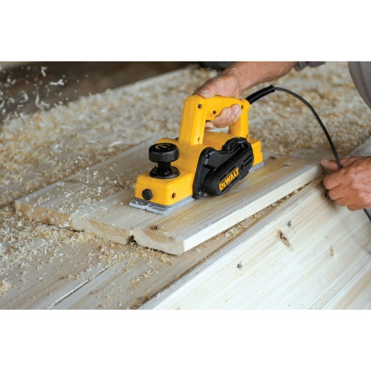 A person using a yellow DEWALT electric hand planer to smooth wooden boards, with wood shavings scattered around the workspace.