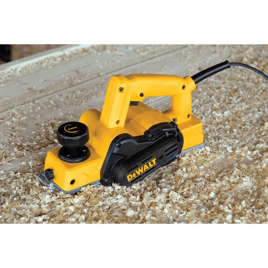 A yellow and black DEWALT electric hand planer is shown in use on a wooden work surface, surrounded by wood shavings.