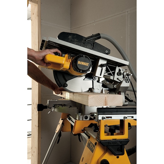 A person is using a DEWALT D27113 saw to cut a piece of wood. The saw is mounted on a yellow stand and is operating in an indoor workshop.