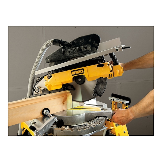A person is using a DEWALT compound miter saw (model D27113) to cut a piece of wood. The saw is yellow and mounted on a workbench.