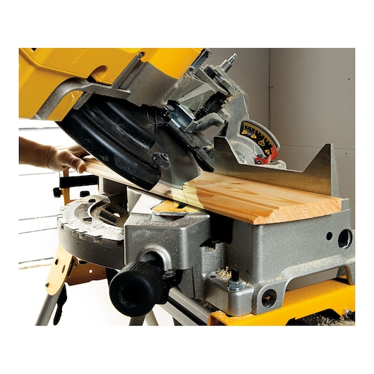 A close-up of a DEWALT miter saw cutting a piece of wood at an angle. The saw is mounted on a workbench, and a person's hand is holding the wood piece in place. Sawdust is visible on the surface.