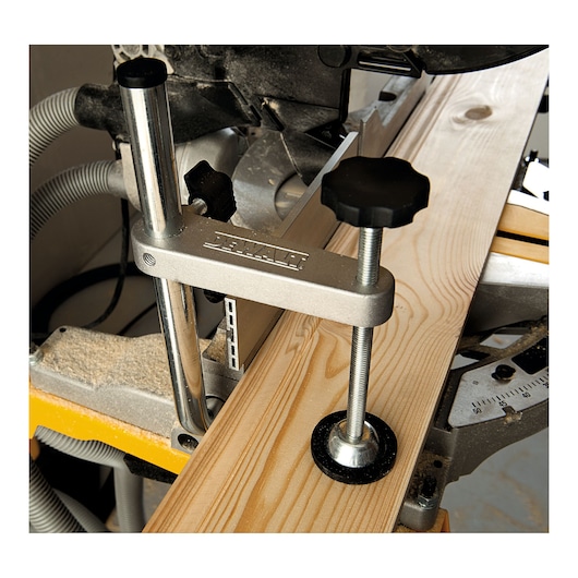 Close-up of a DEWALT clamping system holding a wooden board securely on a miter saw, with adjustment knobs and metal bars visible.