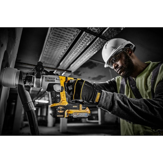 A person wearing construction gear, including gloves and a helmet, is using a DEWALT cordless power tool with a DCBP034 18V