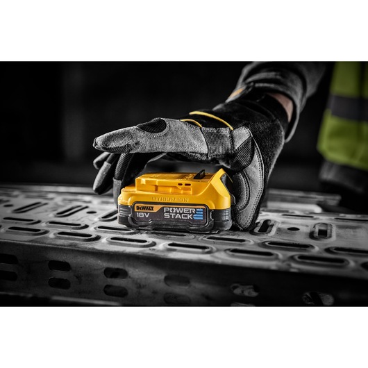 Gloved hand holding a DEWALT 18V POWERSTACK battery on a metal surface at a worksite.