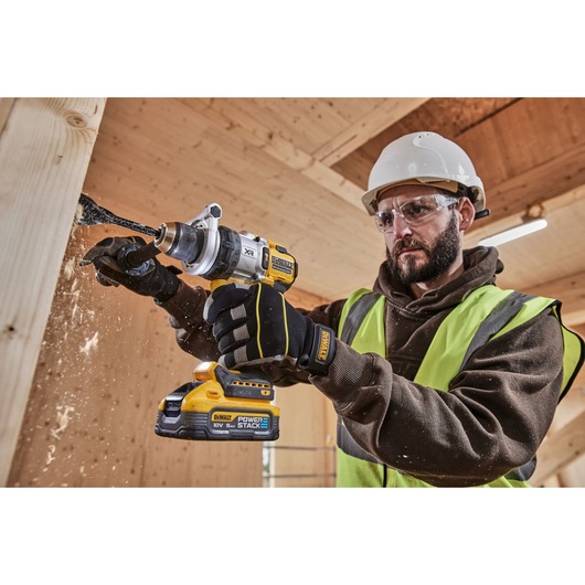 A person wearing a helmet and high-visibility vest is using a DEWALT cordless drill to drill into a wooden beam. Wood shavings are being produced as the drill bit penetrates the surface. The DEWALT drill features a POWERSTACK 18V battery.