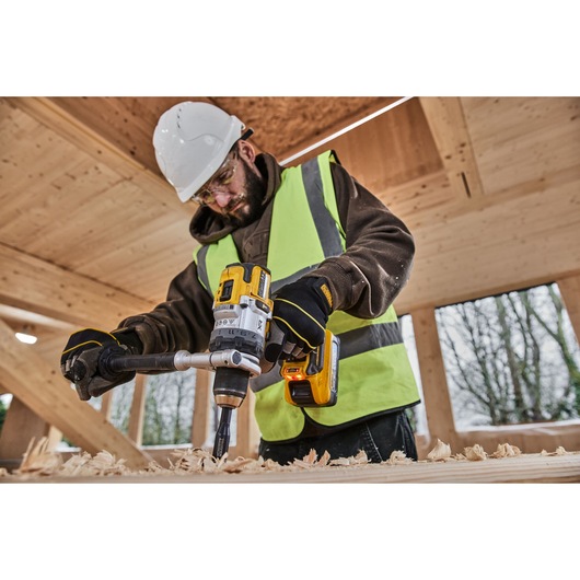 A person wearing a hard hat, high-visibility vest, and gloves is using a DEWALT cordless drill (SKU: DCD1007NT) to drill into a wooden surface inside a construction site.