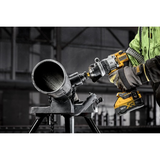 A close-up of a person wearing safety gloves and a jacket using a DEWALT cordless power drill to work on a large metal pipe in an industrial setting.