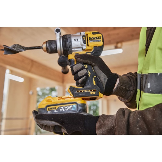 Close-up view of hands wearing gloves holding a DEWALT cordless drill with a POWERSTACK 18V 5Ah battery attached, being used in a construction setting.