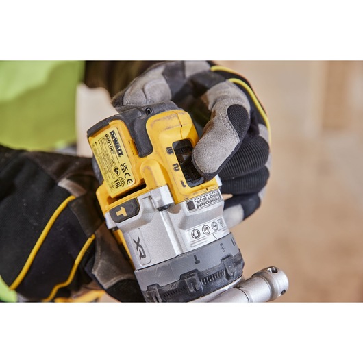 A close-up of a person wearing work gloves adjusting the settings on a DEWALT cordless power tool. The tool features a yellow and silver design with visible labels and controls.