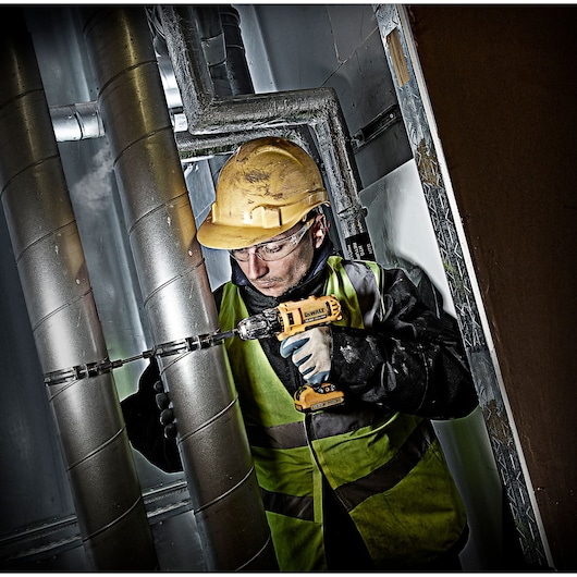 A construction worker wearing a yellow hard hat and safety vest is using a DEWALT cordless drill to work on metal pipes in an industrial setting. The worker's face is blurred for privacy.