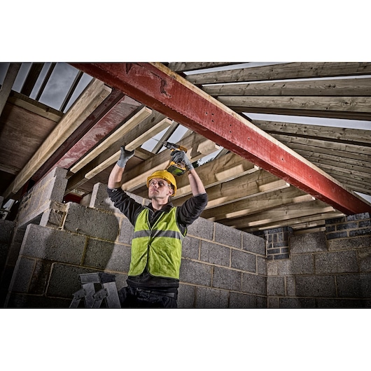 A construction worker wearing a safety vest and helmet uses a DEWALT cordless drill to secure wooden beams in place inside a partially built structure with exposed brick and wooden framework.