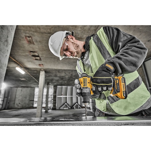 A person wearing a hard hat, safety vest, and gloves is using a DEWALT DCD800D2T cordless drill at a construction site, drilling into a metal surface.