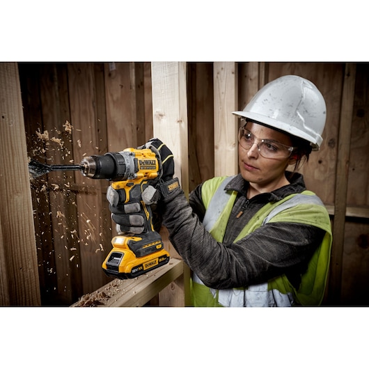 A person wearing safety gear and gloves is using a DEWALT DCD800D2T cordless drill to bore a hole into a wooden beam. Wood chips are flying as the drill operates. The person's face is intentionally blurred for privacy.