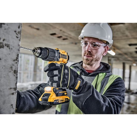 A construction worker wearing safety gloves, a high-visibility vest, and a hard hat is using a DEWALT cordless drill to drill into a concrete surface on a construction site.