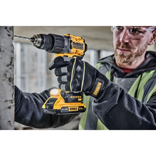 A person wearing work gloves is using a yellow and black DEWALT cordless drill (model DCD805) to drill into a concrete surface. The drill shows the 18V XR 2.0Ah battery attached.
