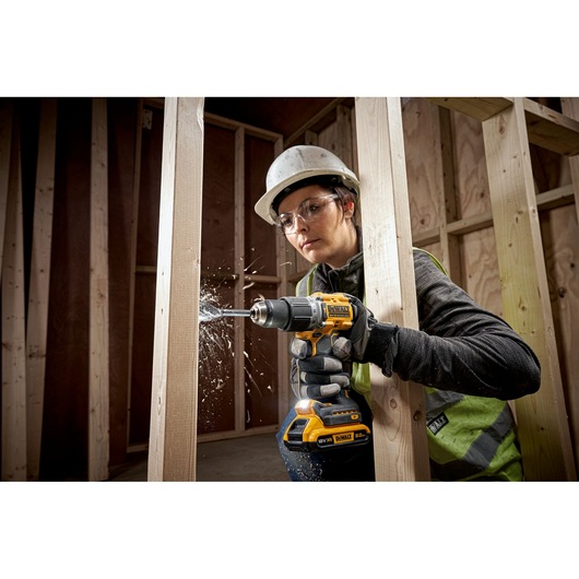A person wearing a hard hat and safety vest uses a DEWALT cordless drill to bore into a wooden beam at a construction site. Wood shavings are flying from the drilling process.