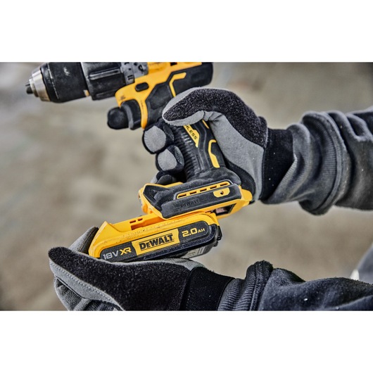 A close-up view of a person wearing gloves inserting an 18V XR 2.0Ah DEWALT battery into a yellow DEWALT cordless power drill.