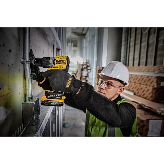 A person wearing protective gear and a safety helmet uses a DEWALT DCD805E2T cordless power drill to work on a metal pipe attached to a wall inside an industrial or construction site.