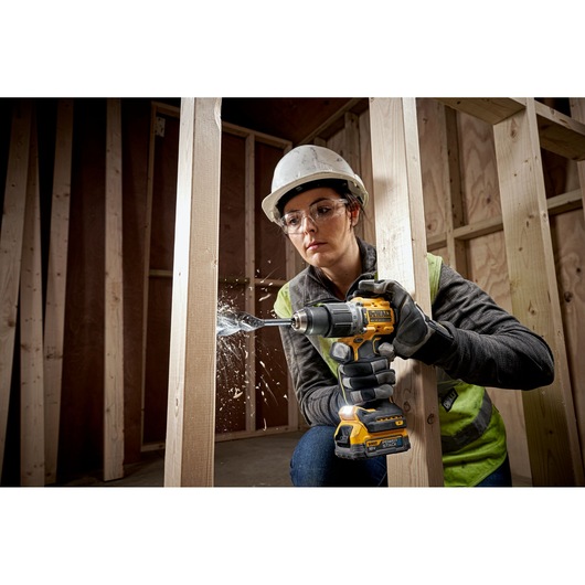 A person wearing a hard hat and safety vest is using a DEWALT DCD805E2T cordless drill to drill into a wooden beam at a construction site. Wood shavings are visible flying from the drill bit.
