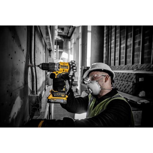 A worker wearing a safety helmet and gloves is using a yellow DEWALT DCD805E2T cordless drill to fasten a metal pipe in an industrial construction setting.
