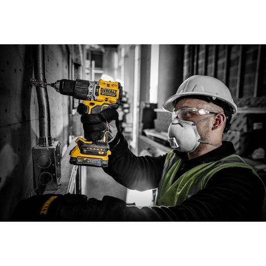 A person wearing a safety helmet and high-visibility vest is using a DEWALT DCD805E2T cordless drill to work on a metal pipe attached to a wall in an industrial setting. The person's face is blurred for privacy.