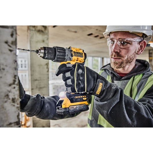 A person wearing safety gloves and a safety vest is using a DEWALT 18V cordless drill to drill into a metal surface at a construction site. The drill is branded DEWALT and fitted with a POWERSTACK battery.
