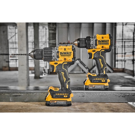 Two DEWALT cordless power drills, model DCD805NT, are positioned side by side on a construction site workbench. Both drills are yellow and black with mounted battery packs labeled 'POWERSTACK 18V'. One drill has a metal drill bit attached.