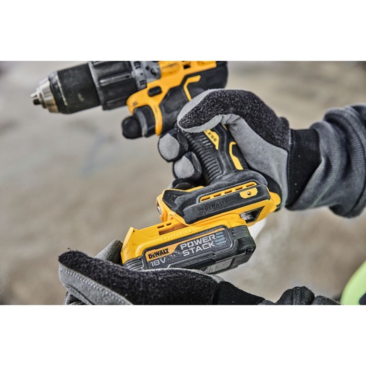 Close-up of gloved hands holding a yellow and black DEWALT cordless drill, with visible 18V POWERSTACK battery being attached.