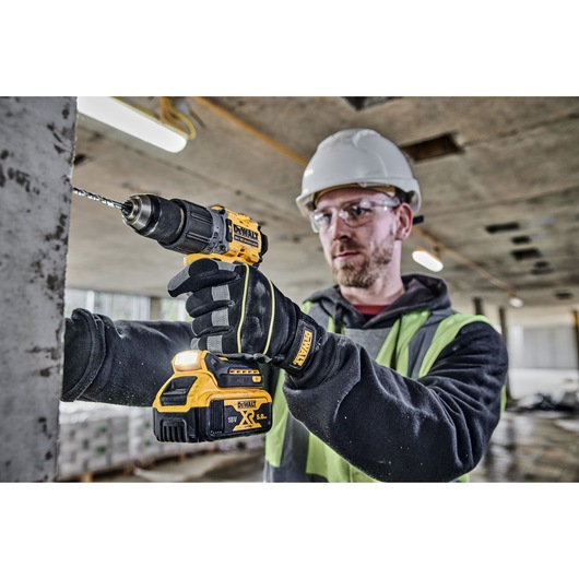 A person wearing gloves, a safety vest, and a hard hat is using a DEWALT DCD805P2T cordless drill to drill into a concrete surface at a construction site. The face is blurred for privacy.