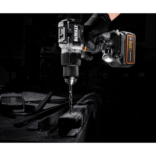 A DEWALT cordless drill is being used to drill into a metal surface. The drill is held by a gloved hand and is equipped with a battery pack. The scene is well-lit, highlighting the drill and the work area.