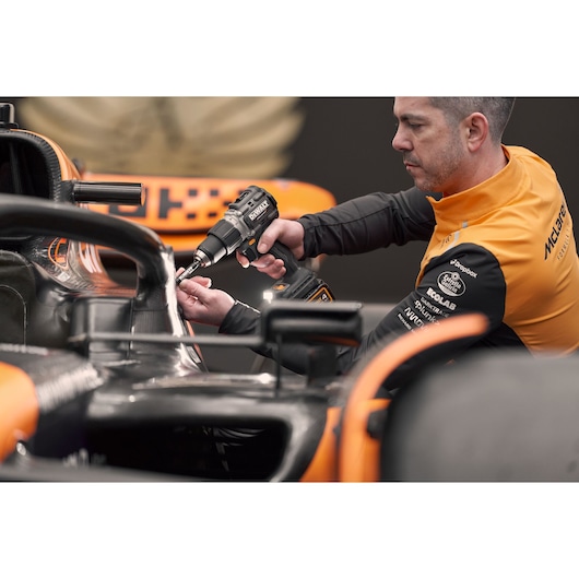A person with a blurred face uses a DEWALT cordless drill to work on a part of an orange and black racing car. The person is wearing an orange and black jacket with various sponsor logos visible.