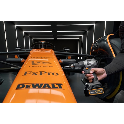 A close-up view of an orange Formula 1 race car with various sponsor logos, including DEWALT, as a person uses a DEWALT cordless drill on the car in a modern, illuminated garage.