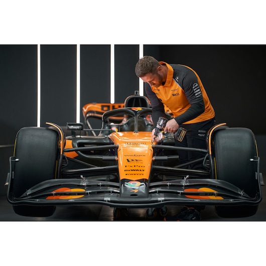 A person with their face blurred is using a DEWALT power tool on the front section of an orange Formula 1 race car inside a garage or workshop setting.