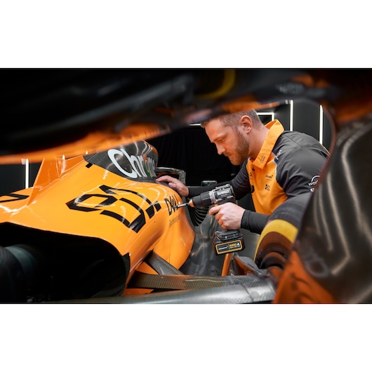A person wearing an orange and black uniform is using a DEWALT cordless drill on the side of an orange race car. The person's face is blurred and the DEWALT branding is visible on the tool and the car.
