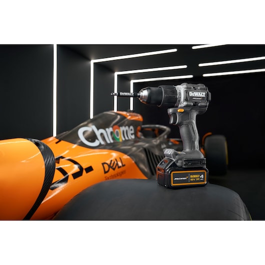 A DEWALT cordless drill with a drill bit attached is placed on a car tire in front of an orange and black racing car, with sponsor logos visible. The drill battery displays both DEWALT and McLaren branding. The background features dark walls with illuminated white light strips.