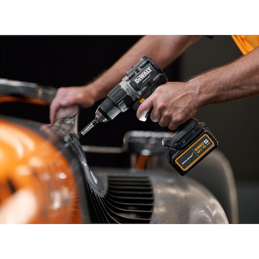 A close-up of a person using a DEWALT cordless drill with a battery pack to work on a metallic object, possibly an automobile part.